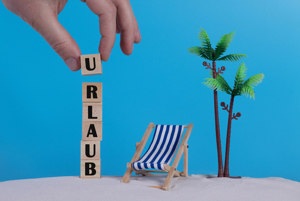 Hand with wooden blocks wiht Urlaub text and beach chair on blue background Hand with wooden blocks wiht Urlaub text and beach chair on blue background