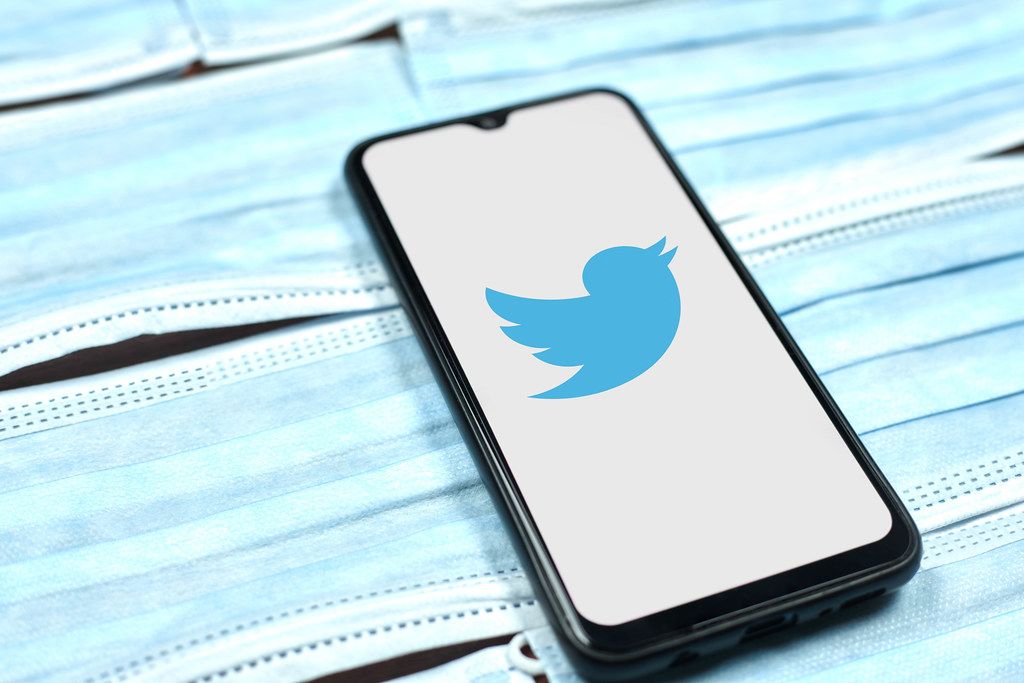 Twitter logo on smartphone screen over the face masks. Global company during coronavirus crisis Twitter logo on smartphone screen over the face masks. Global company during coronavirus crisis