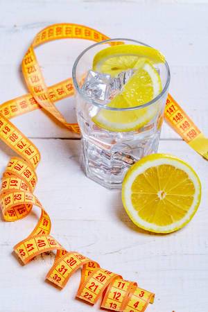 Glass of cold water with ice cubes and lemon on white wooden background with measuring tape-healthy lifestyle concept Glass of cold water with ice cubes and lemon on white wooden background with measuring tape-healthy lifestyle concept